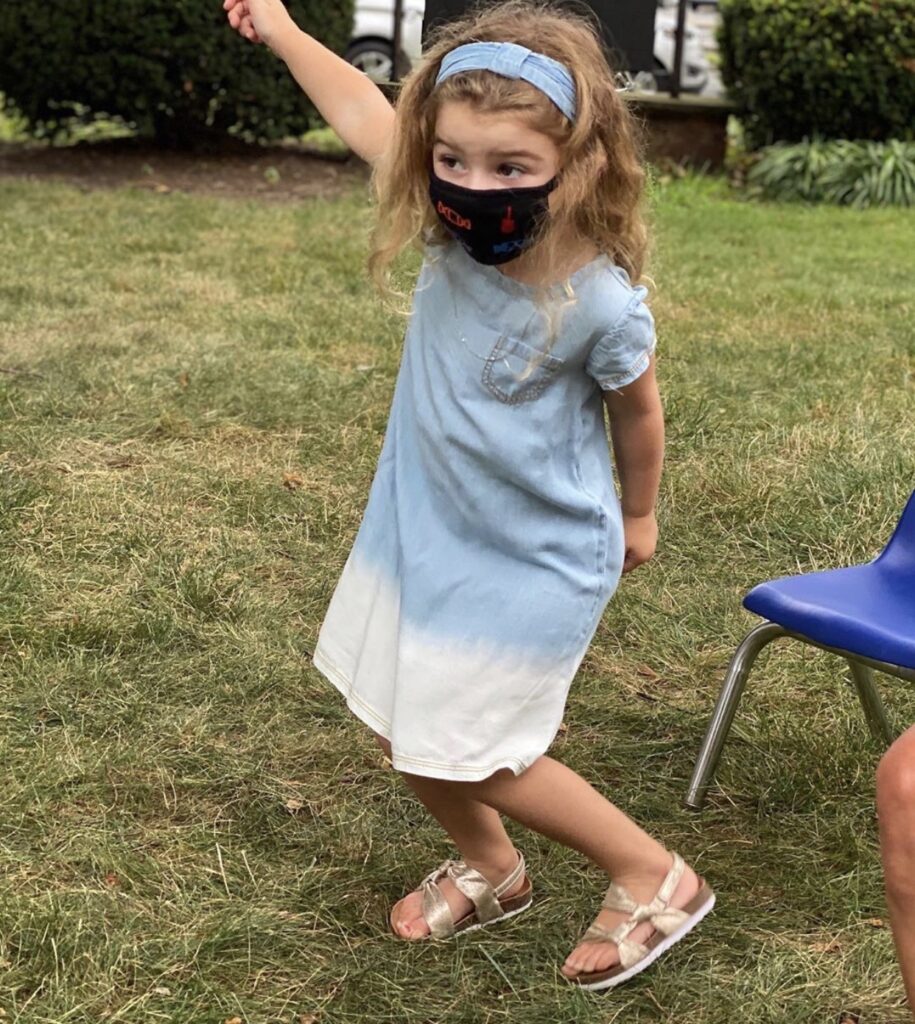 Young girl in a blue and white dress wearing a black mask outdoors.